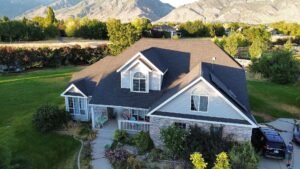 Drone view of a two-story Highland, Utah home with a dark architectural shingle roof, two front gables with dormer windows and the Wasatch Mountains behind it.