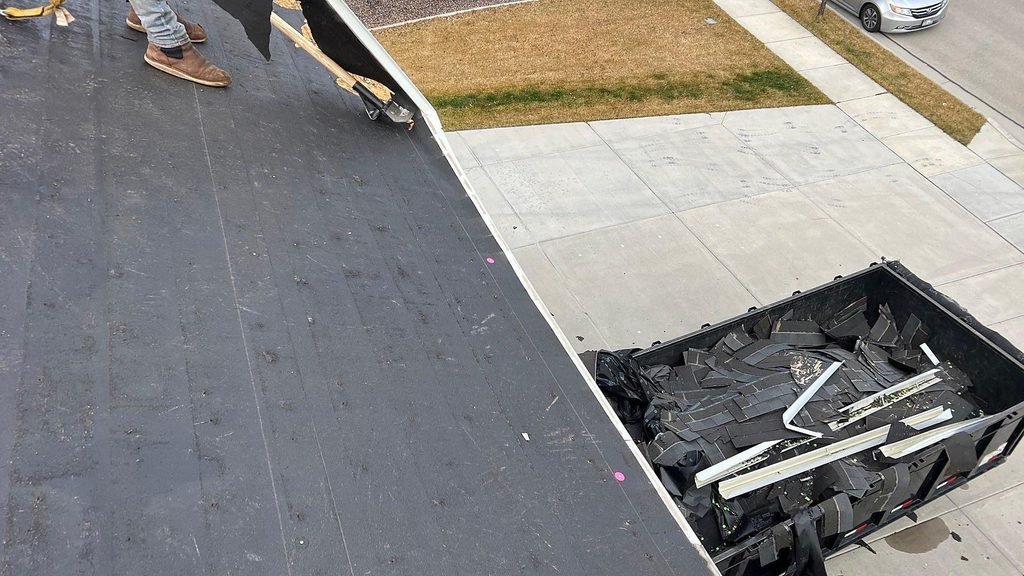 View from a roof in Highland, Utah, looking down at a dump trailer full of torn-off shingles parked at the eave beside the driveway.
