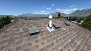 Weathered architectural asphalt shingle roof with box vents and a vent pipe, with the snow-capped Wasatch Mountains behind