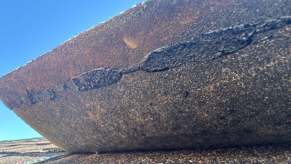 Close-up of an aging asphalt shingle cracking along its edge as it is lifted