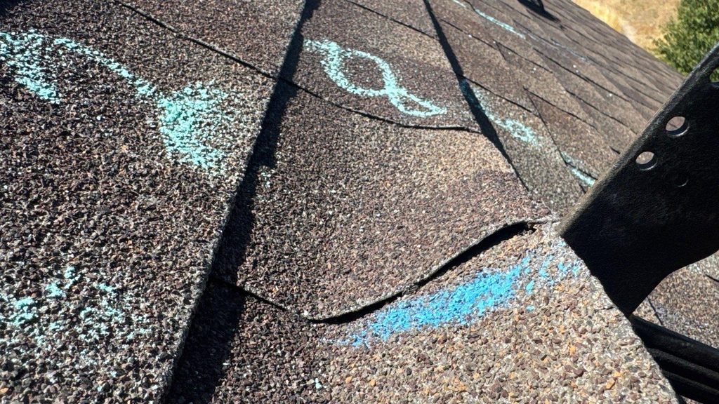 Creased asphalt shingle tab with a horizontal fold line, marked in blue chalk during a roof inspection