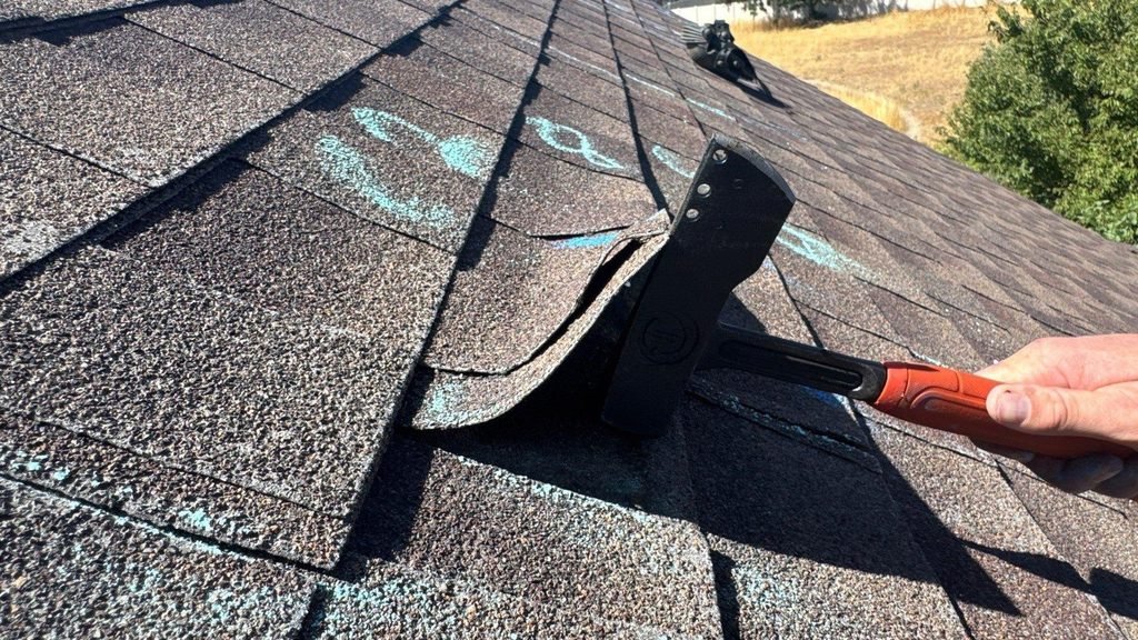 Roofer lifting an asphalt shingle tab with a flat bar to check whether it is still sealed to the shingle below